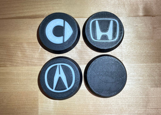 Four round black car emblem covers featuring the logos of Cheetah, Honda, Acura, and a blank black emblem, arranged in a 2x2 grid on a wooden tabletop.
